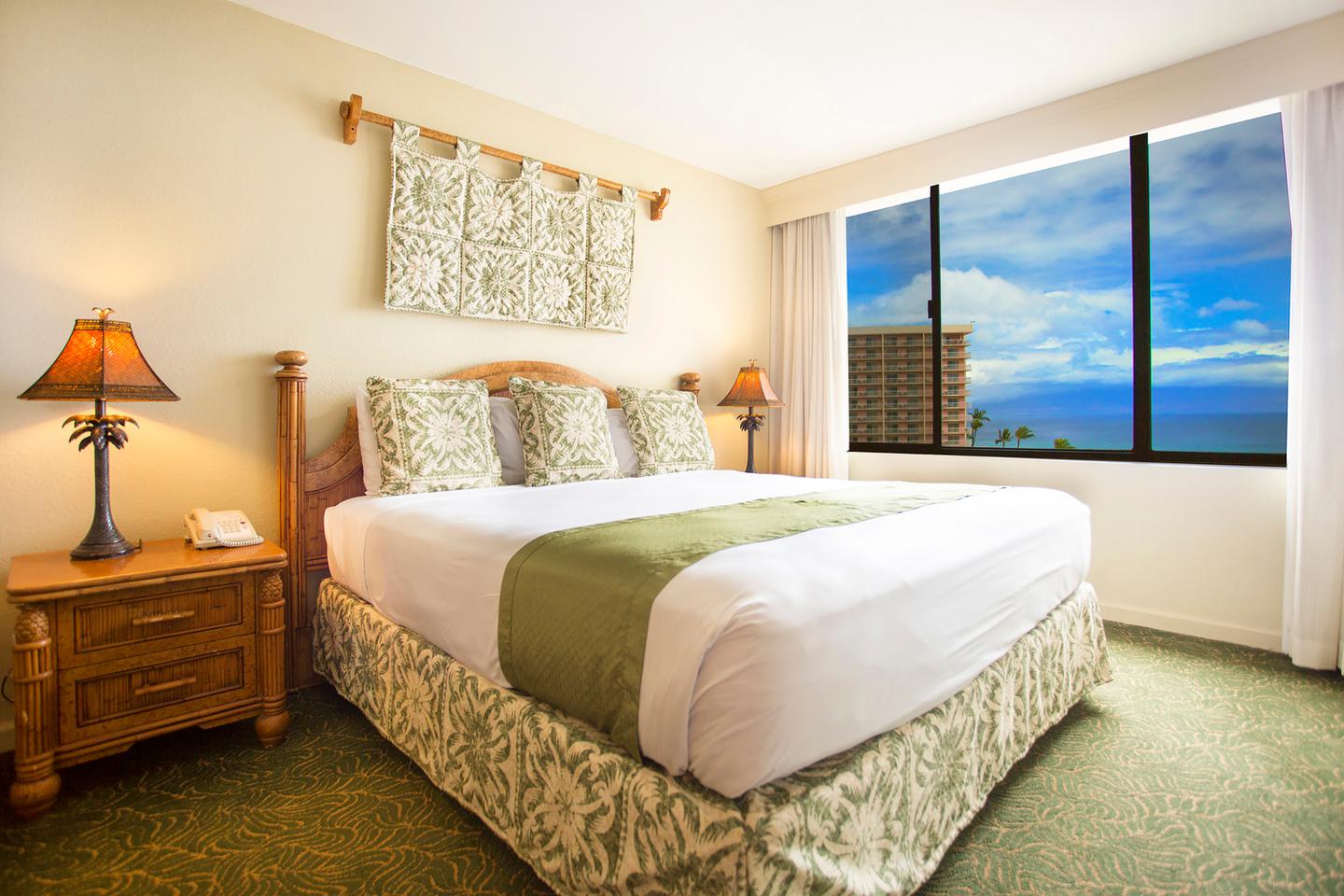 Room with a bed, nightstand, lamps, and picture window with ocean view.