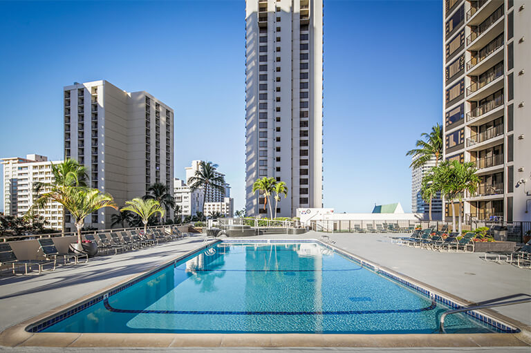 Pool deck at the Aston Waikiki Banyan with deck chairs, palm trees and a separate jet spa
