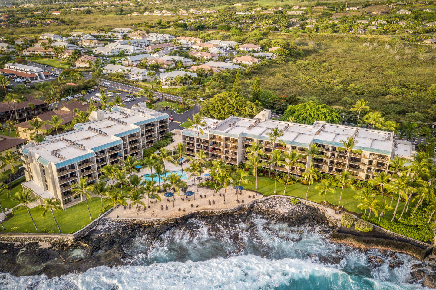 Aerial view of Aston Kona by the Sea lined by black lava rocks, coconut tress, and blue ocean.