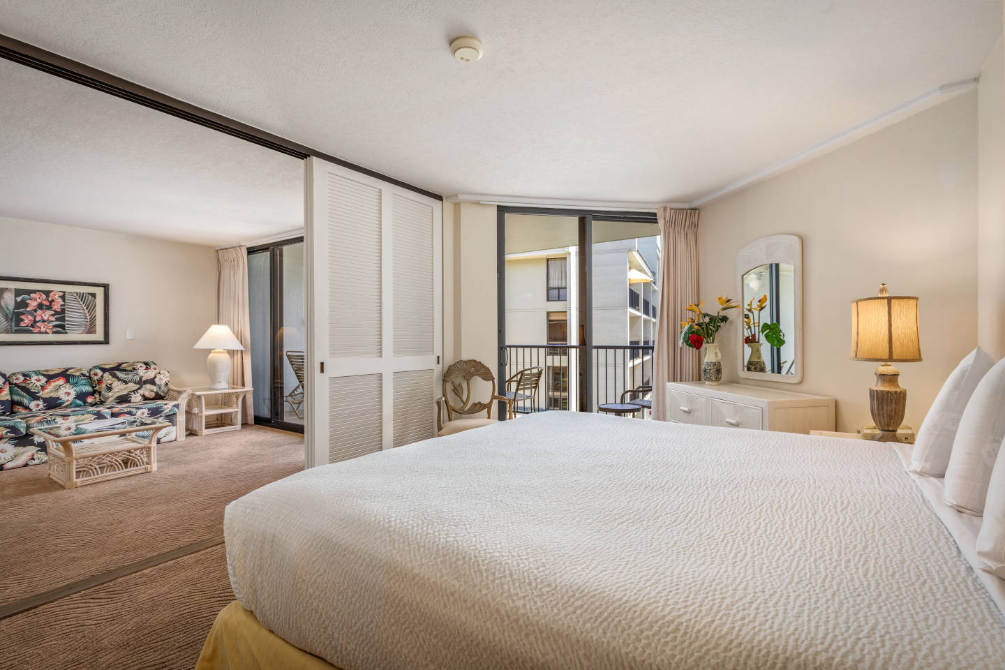 Room with king bed, dresser, side table, lamp,  balcony overlooking the pool area and sliding doors to living room.