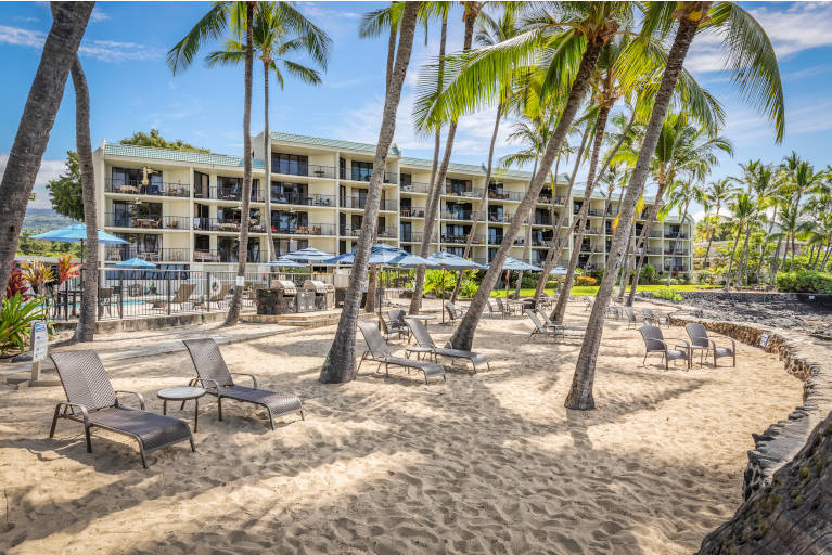 Sand area with lounge chairs near the shoreline with the hotel in the background shaded by coconut trees