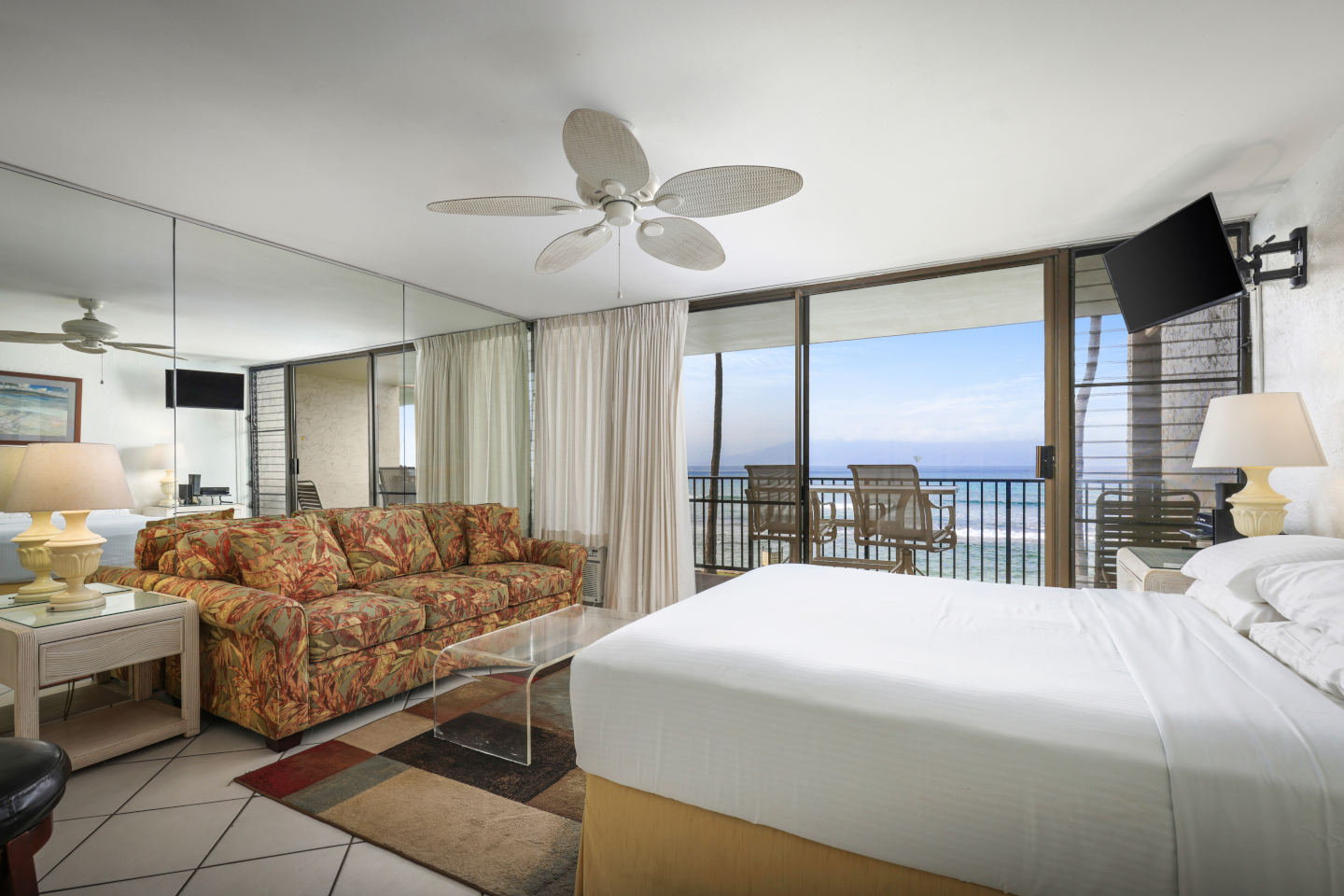 Room with a bed, night stand, lamps, couch, TV, and balcony wth ocean view.