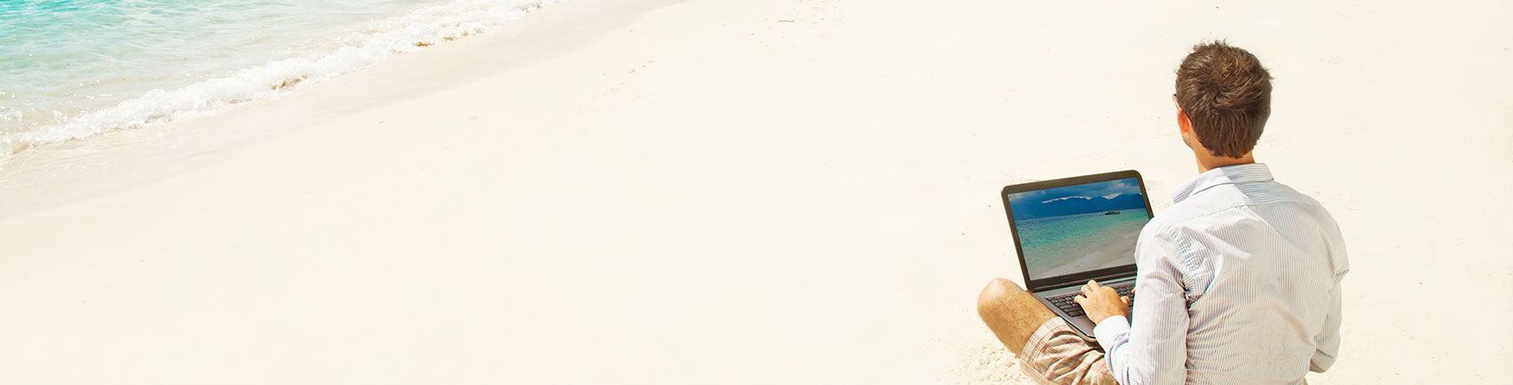 Man sitting crossed legged on the sand in a long sleeve shirt with a laptop in his lap.