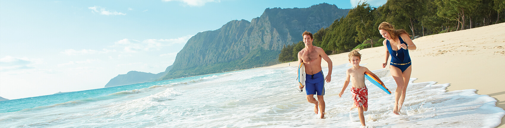 Woman and man with a young boy carrying boggie boards running along the white sand beach
